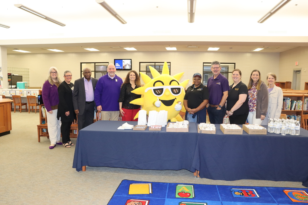 A group of administrators and staff stand together behind a table of breakfast items in the media center, posing with a large yellow sun mascot in the center.