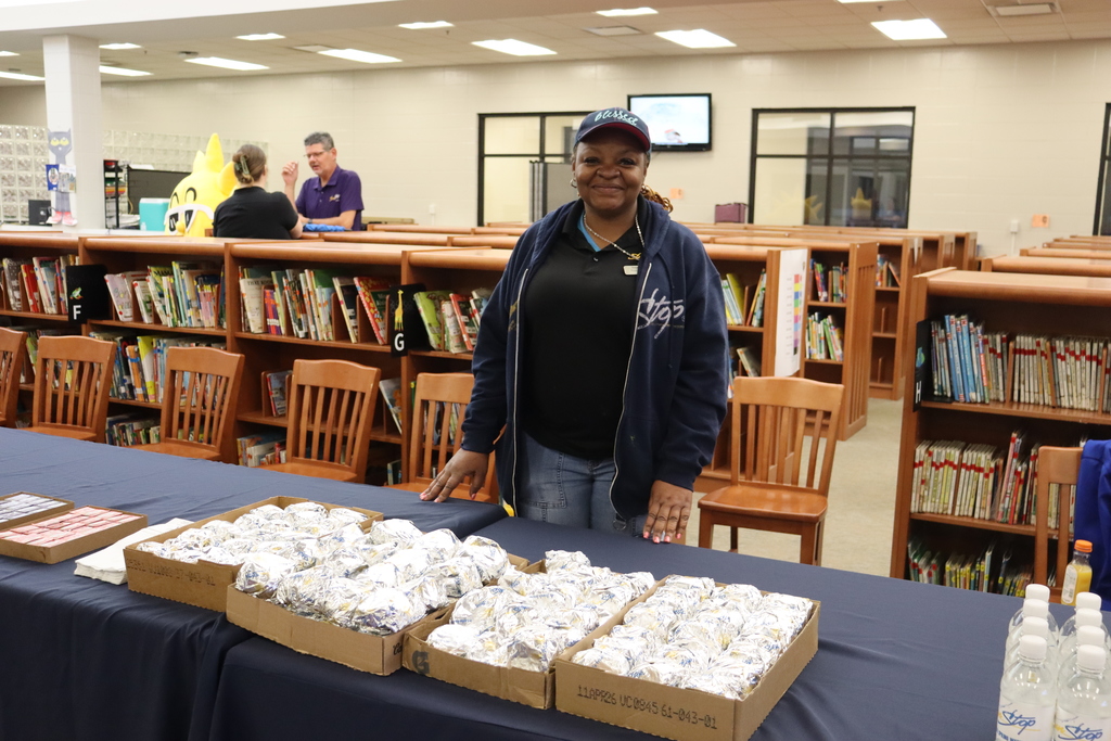A staff member stands behind a table covered with boxed, wrapped breakfast biscuits and bottled drinks inside a school library, with bookshelves and other staff in the background.