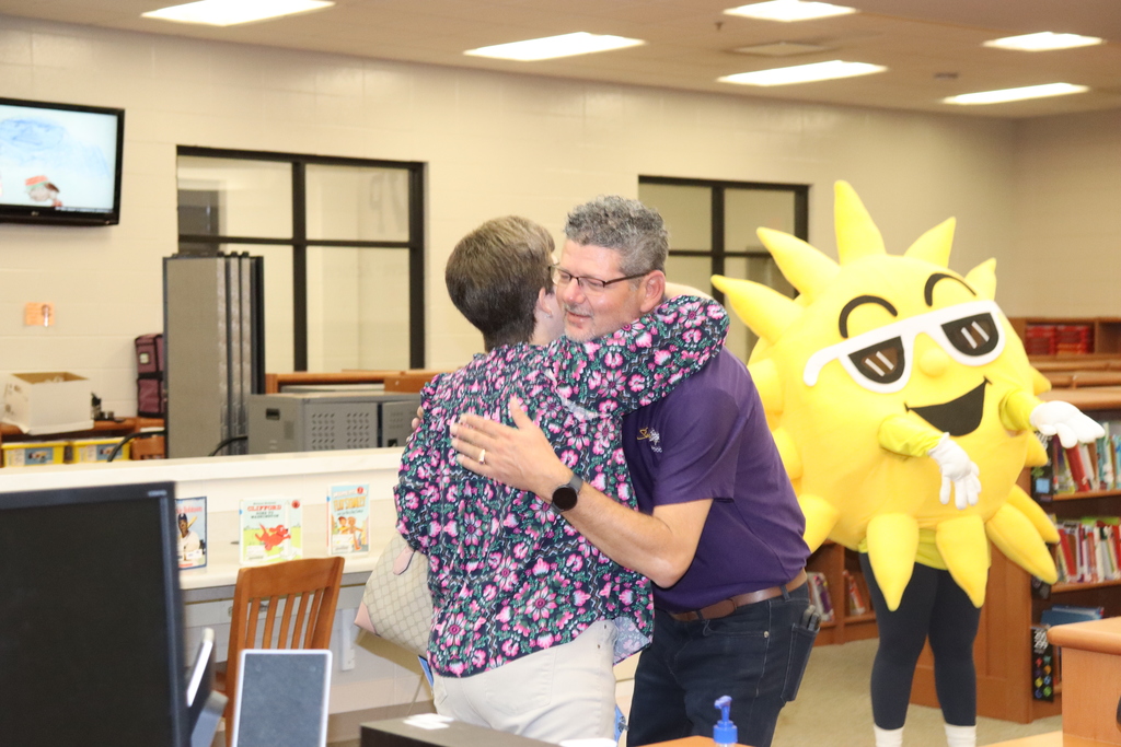 A man in a purple shirt hugs a staff member in the media center while a bright yellow sun mascot wearing sunglasses stands nearby between the bookshelves.