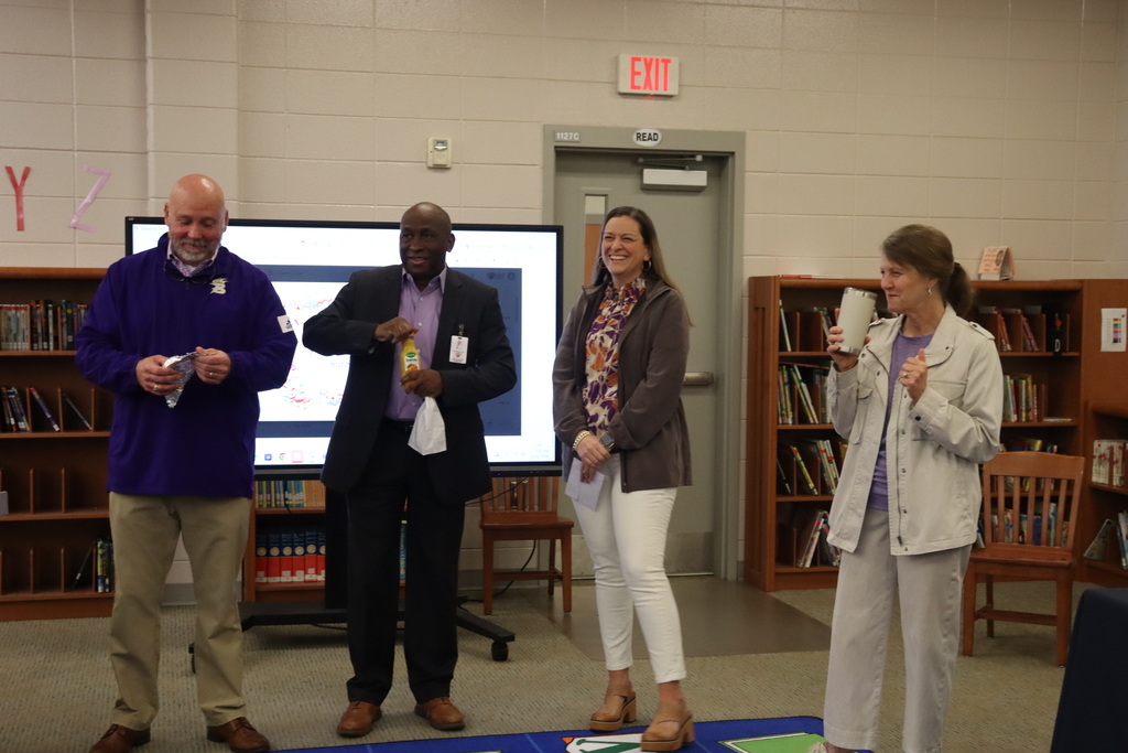 Four administrators stand at the front of the media center smiling and opening breakfast items while a presentation screen is visible behind them.