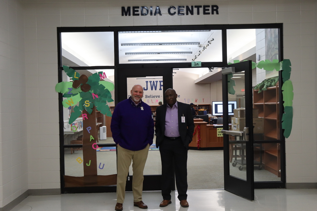 Two administrators stand smiling in the doorway of a school media center decorated with a paper tree and colorful letters spelling “LMNOP…READ.” The sign above the entrance reads “MEDIA CENTER.”