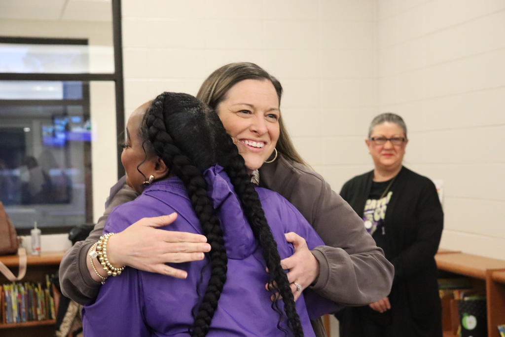 Two women share a hug in the media center while another staff member looks on, smiling, with bookshelves visible behind them.