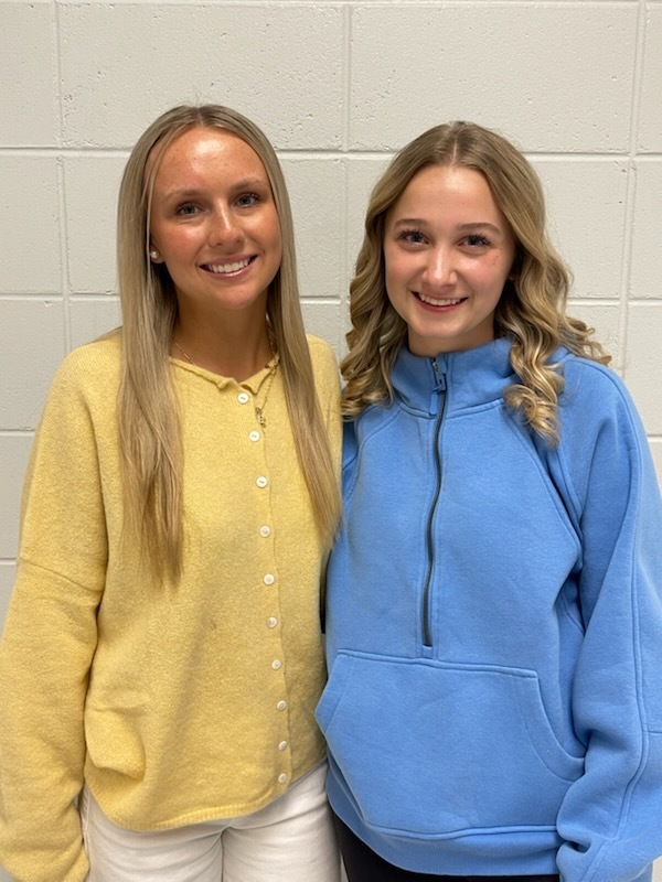 Two young women smiling and standing side by side in front of a white brick wall, one wearing a yellow cardigan and the other a blue quarter-zip pullover.