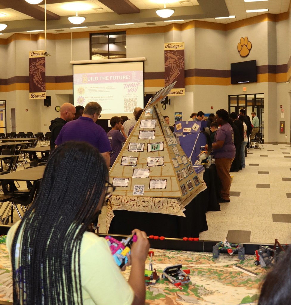 Students and adults explore multiple robotics project displays set up throughout the cafeteria, including tri-fold boards and competition tables, during a school STEM showcase event.
