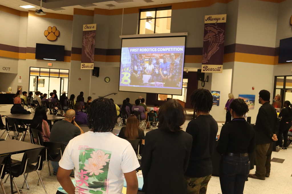 Students and adults sit at cafeteria tables watching a presentation projected on a large screen titled “FIRST Robotics Competition,” inside a school cafeteria decorated with Bearcat banners and school colors.