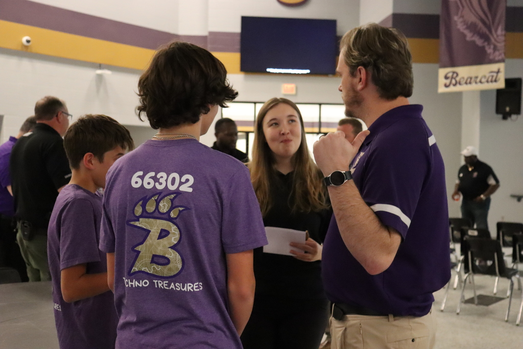A group of middle school students wearing purple team shirts talk with an adult male in a purple polo inside the school cafeteria, discussing their robotics team project.
