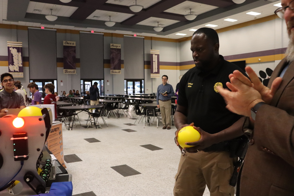 A school resource officer and another adult watch a robotics demonstration, with the officer holding a yellow foam ball while observing a student-built robot display.