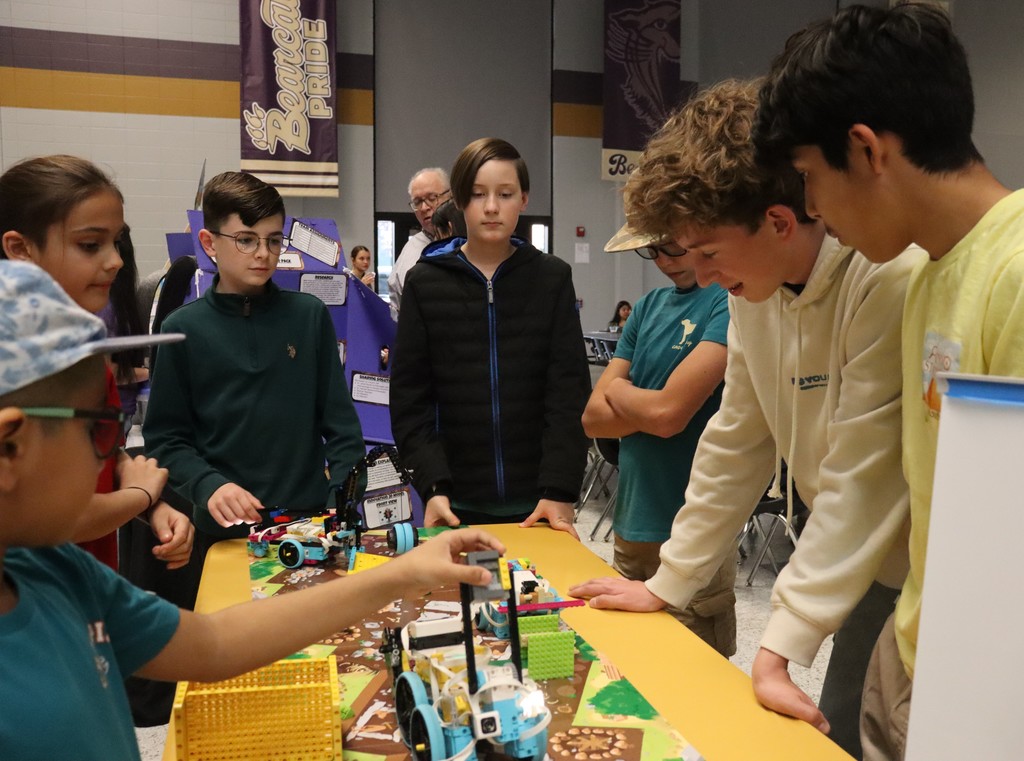 Several students gather around a robotics competition table, closely examining and discussing their LEGO-style robots and competition mat in the school cafeteria.