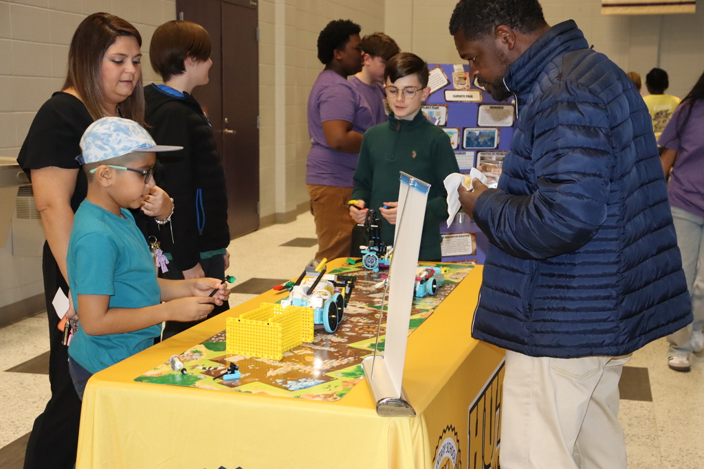 Students present their robotics build to two adults at a display table covered with a yellow cloth, explaining how their LEGO-based robot works during a school showcase event.