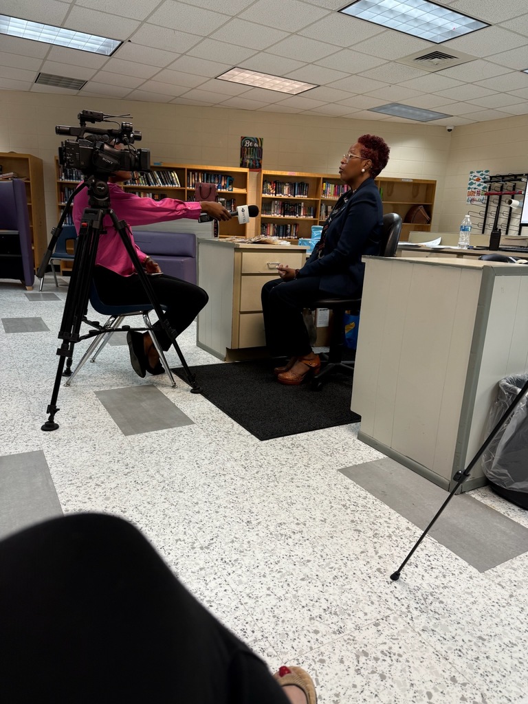 A TV news reporter interviews a staff member seated at a desk in a school library, holding a microphone while a camera records the conversation.