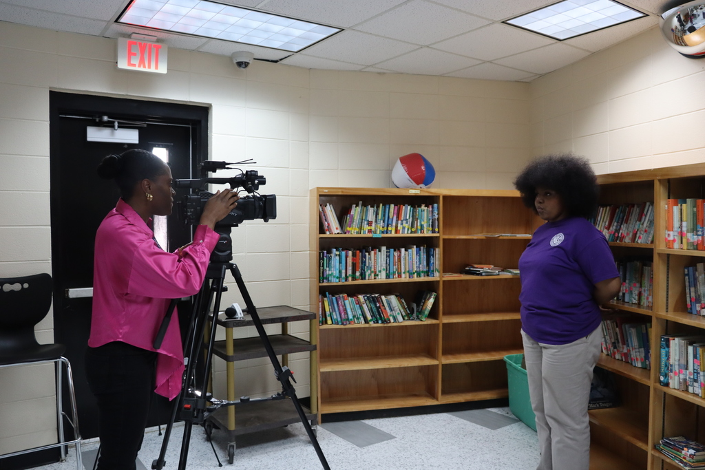 A TV news reporter films a student standing in a school library, with bookshelves in the background, as part of an interview segment.