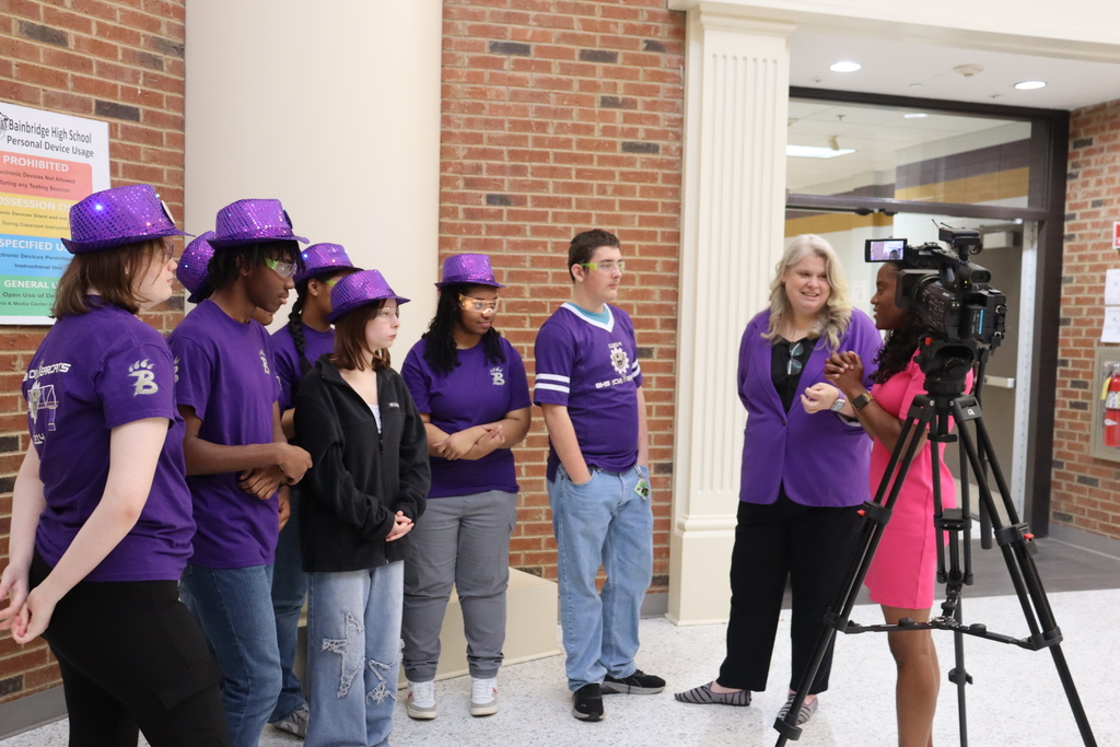 Bainbridge High School robotics students in purple shirts and hats are interviewed on camera by a reporter inside the school.