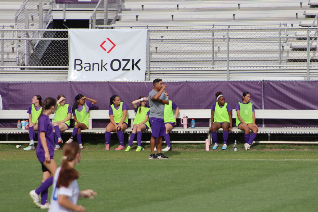 Bainbridge High School Lady Cats soccer players compete in a game, with a goalkeeper clearing the ball and teammates in purple advancing against defenders in white on the field.