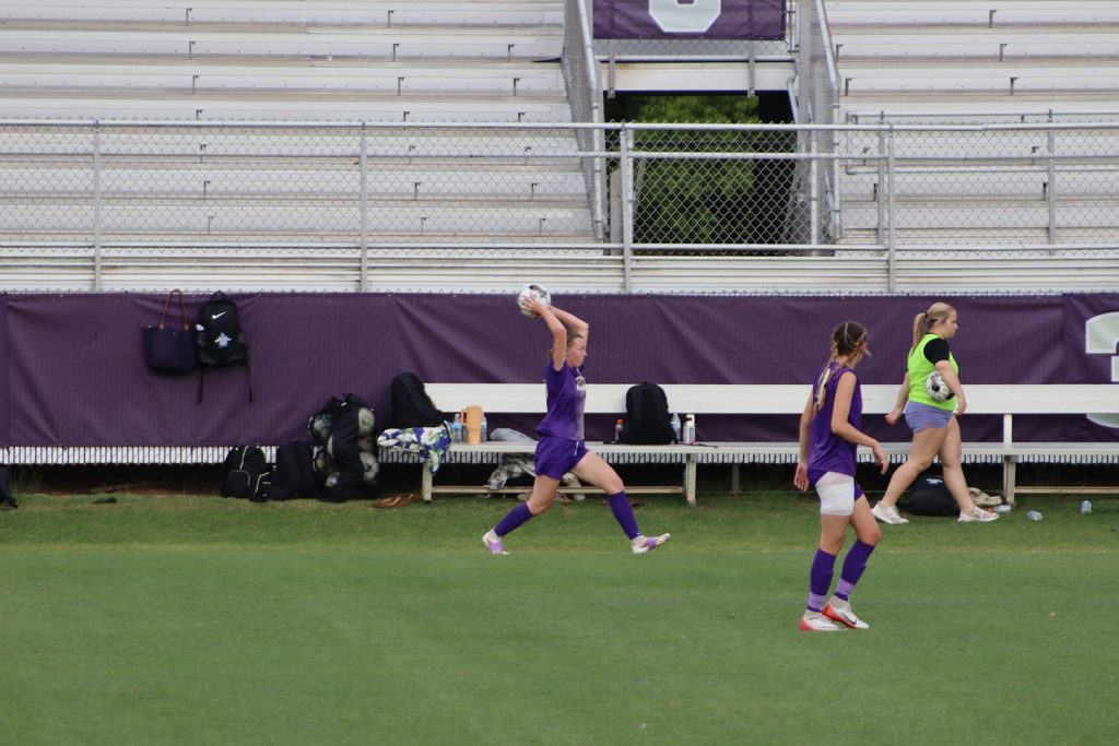 Bainbridge High School Lady Cats soccer players compete in a game, with a goalkeeper clearing the ball and teammates in purple advancing against defenders in white on the field.