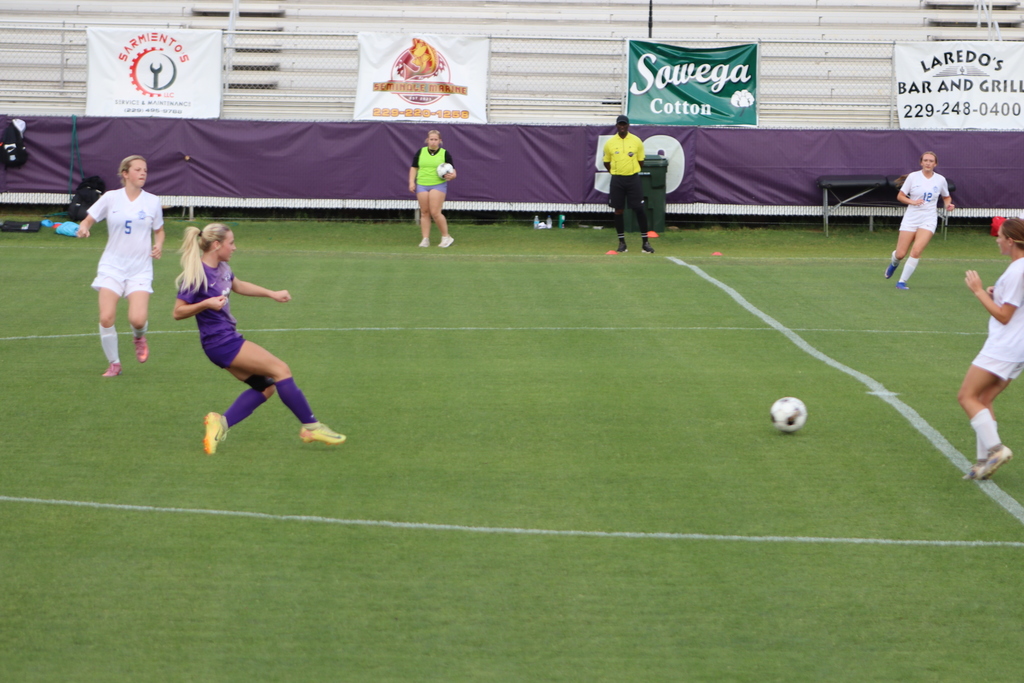 Bainbridge High School Lady Cats soccer players compete in a game, with a goalkeeper clearing the ball and teammates in purple advancing against defenders in white on the field.