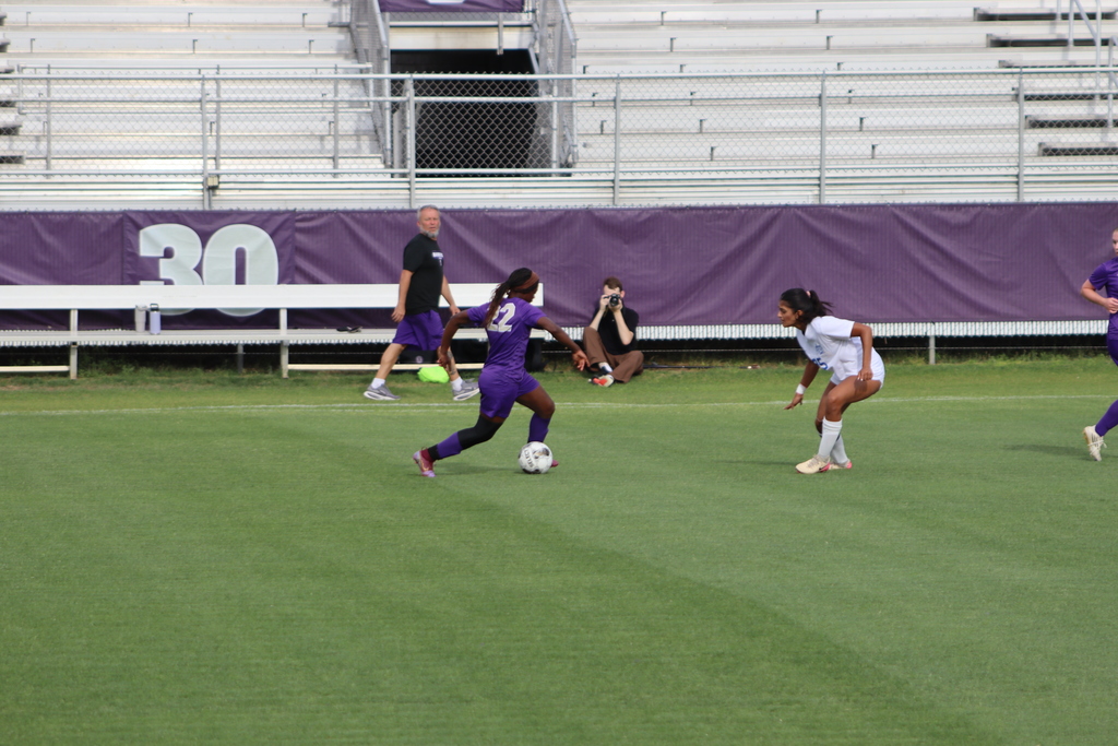 Bainbridge High School Lady Cats soccer players compete in a game, with a goalkeeper clearing the ball and teammates in purple advancing against defenders in white on the field.