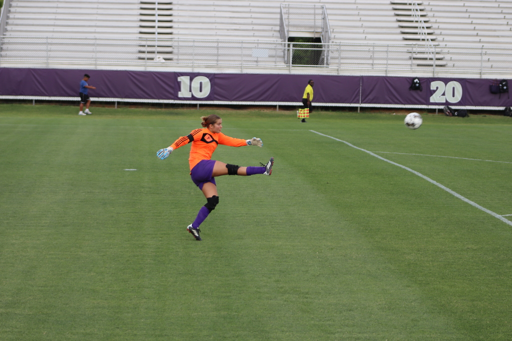 Bainbridge High School Lady Cats soccer players compete in a game, with a goalkeeper clearing the ball and teammates in purple advancing against defenders in white on the field.