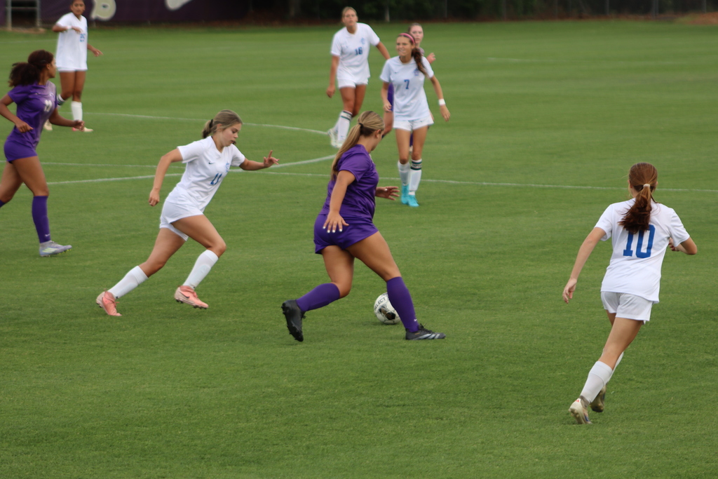Bainbridge High School Lady Cats soccer players compete in a game, with a goalkeeper clearing the ball and teammates in purple advancing against defenders in white on the field.