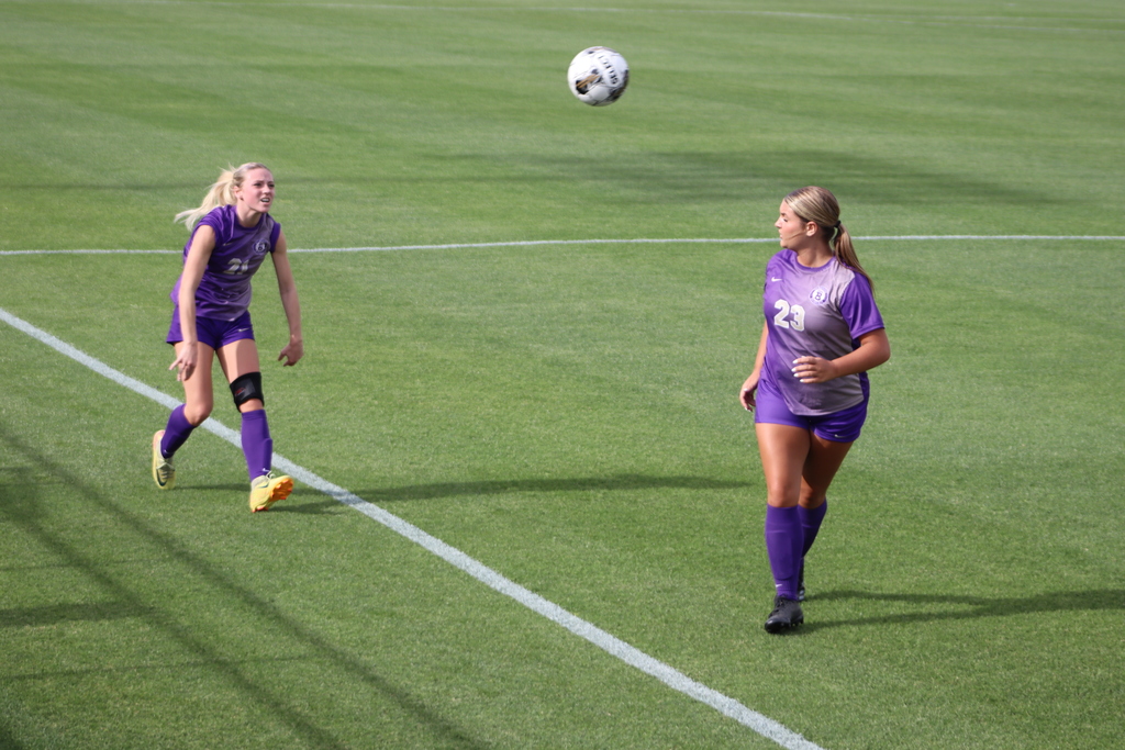 Bainbridge High School Lady Cats soccer players compete in a game, with a goalkeeper clearing the ball and teammates in purple advancing against defenders in white on the field.