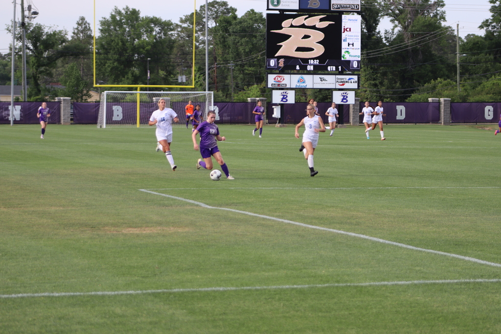 Bainbridge High School Lady Cats soccer players compete in a game, with a goalkeeper clearing the ball and teammates in purple advancing against defenders in white on the field.