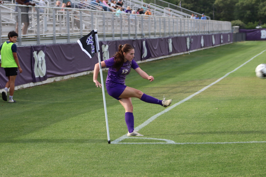 Bainbridge High School Lady Cats soccer players compete in a game, with a goalkeeper clearing the ball and teammates in purple advancing against defenders in white on the field.