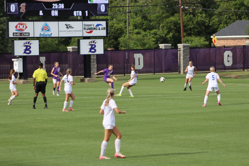Bainbridge High School Lady Cats soccer players compete in a game, with a goalkeeper clearing the ball and teammates in purple advancing against defenders in white on the field.