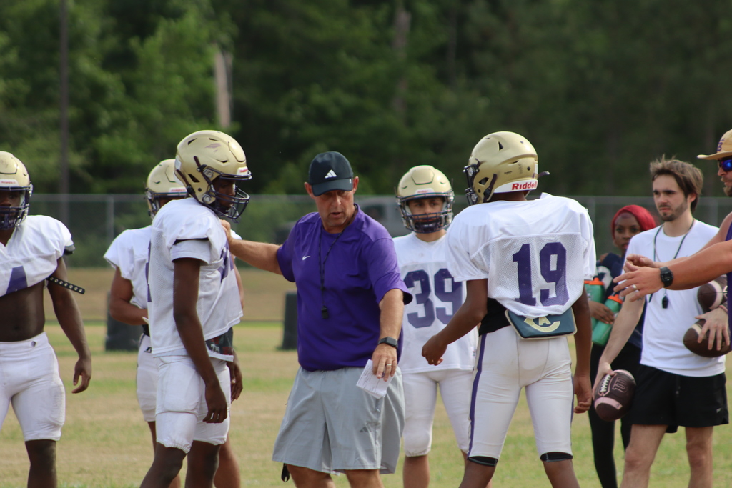 A group of high school football players in purple and white uniforms participate in an intense outdoor practice, running drills, engaging in contact exercises, and listening to coaches on a grassy field surrounded by teammates.