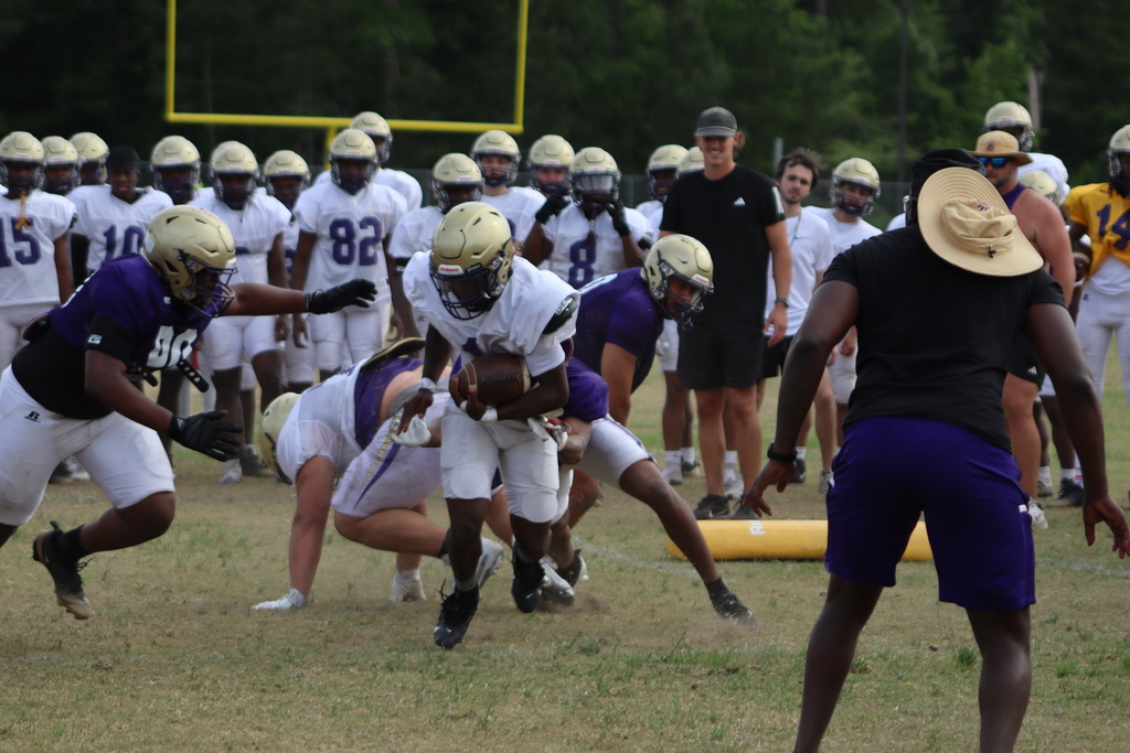 A group of high school football players in purple and white uniforms participate in an intense outdoor practice, running drills, engaging in contact exercises, and listening to coaches on a grassy field surrounded by teammates.