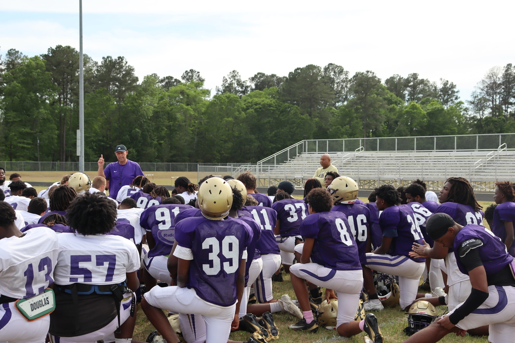 A group of high school football players in purple and white uniforms participate in an intense outdoor practice, running drills, engaging in contact exercises, and listening to coaches on a grassy field surrounded by teammates.