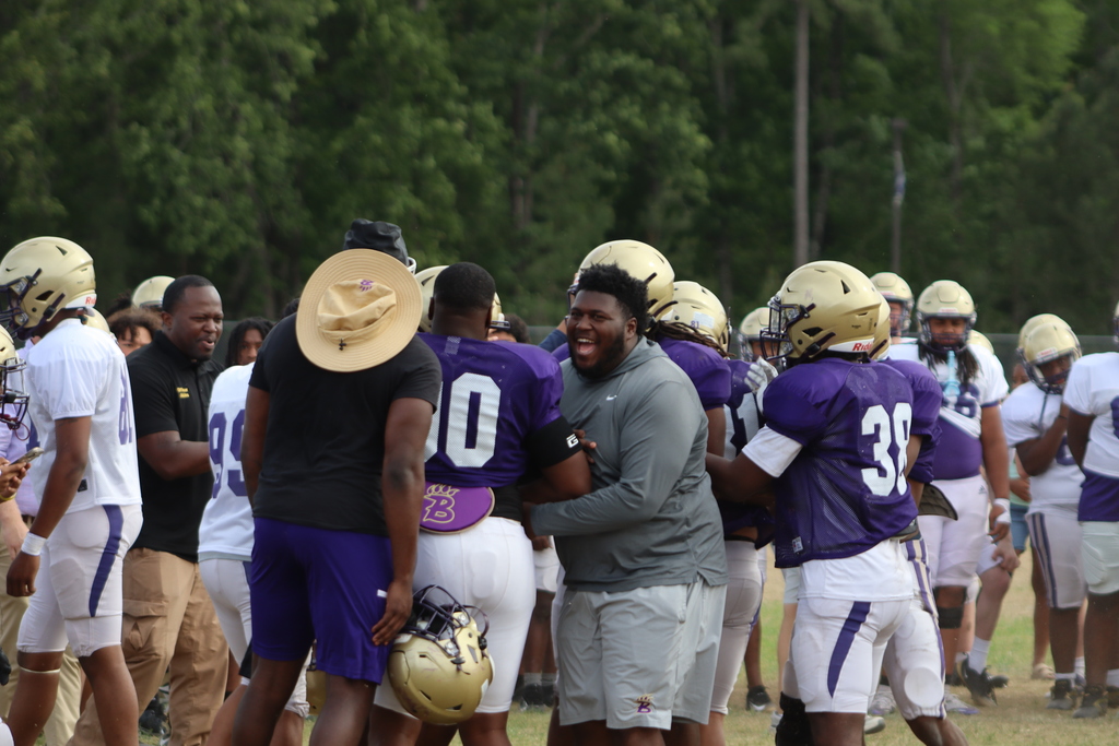 A group of high school football players in purple and white uniforms participate in an intense outdoor practice, running drills, engaging in contact exercises, and listening to coaches on a grassy field surrounded by teammates.