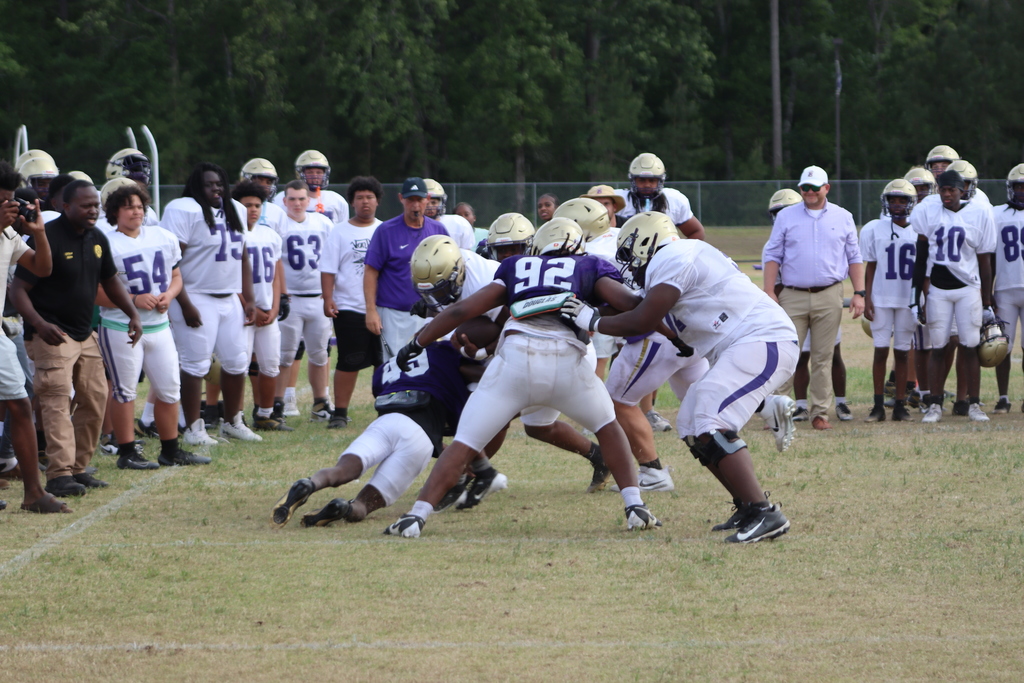 A group of high school football players in purple and white uniforms participate in an intense outdoor practice, running drills, engaging in contact exercises, and listening to coaches on a grassy field surrounded by teammates.