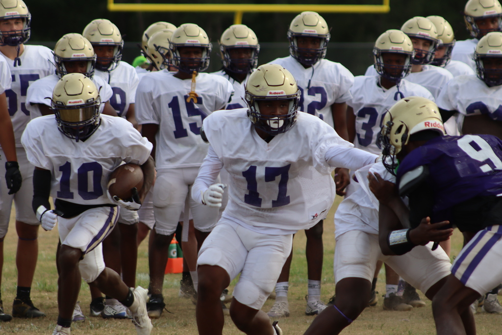 A group of high school football players in purple and white uniforms participate in an intense outdoor practice, running drills, engaging in contact exercises, and listening to coaches on a grassy field surrounded by teammates.