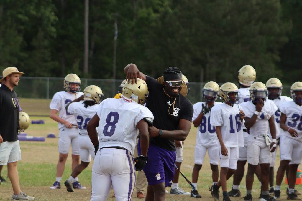 A group of high school football players in purple and white uniforms participate in an intense outdoor practice, running drills, engaging in contact exercises, and listening to coaches on a grassy field surrounded by teammates.