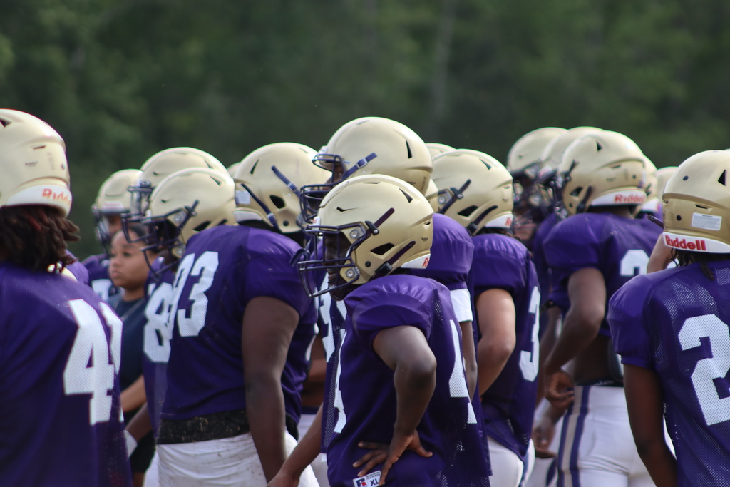 A group of high school football players in purple and white uniforms participate in an intense outdoor practice, running drills, engaging in contact exercises, and listening to coaches on a grassy field surrounded by teammates.