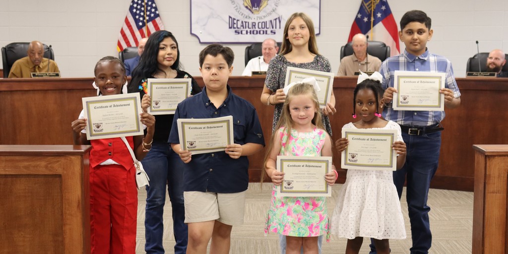 A group of seven elementary-aged students stand in a school board meeting room holding certificates of achievement, posing in front of seated board members. The students are arranged in two rows, smiling and dressed in a mix of casual and semi-formal outfits. Behind them, a wall sign reads “The Bearcat Way – Decatur County Schools,” with the U.S. and Georgia state flags displayed on either side.