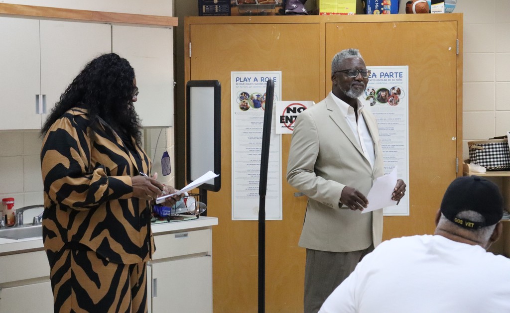 A man and a woman stand at the front of a classroom or meeting space, each holding papers as they address an audience. The man, wearing a light-colored blazer and glasses, faces forward while speaking, and the woman, dressed in a patterned outfit, looks down at her notes. Behind them are wooden doors with educational posters, and a ring light stands between them, suggesting the event may be recorded or photographed. A seated attendee is visible in the foreground.