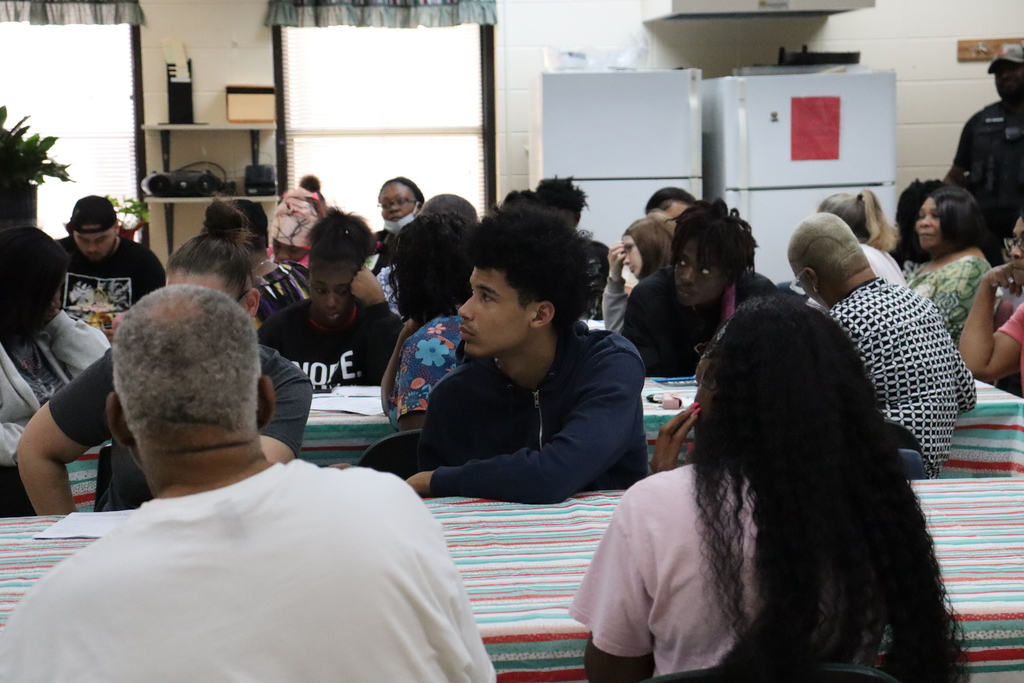 A group of adults seated around tables listen to a woman speaking at the front of a classroom-style meeting room.