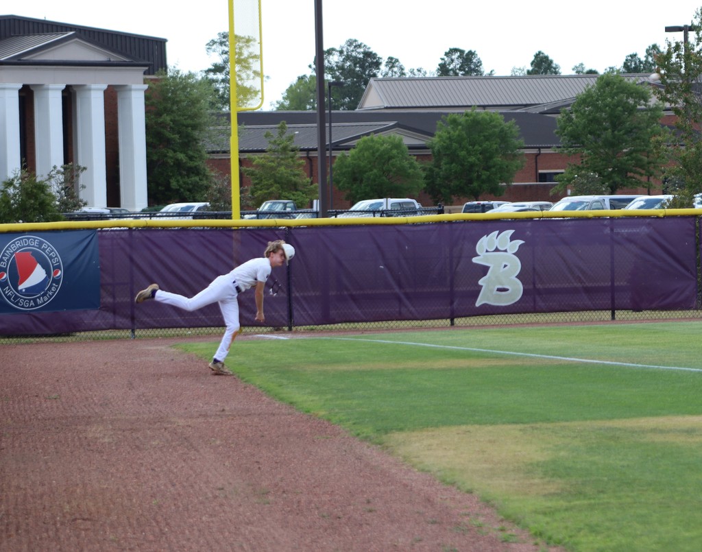 High school baseball game featuring Bainbridge Bearcats players in white uniforms fielding, pitching, catching, and running bases on a dirt and grass field, with a scoreboard, outfield fence signage, and spectators in the background.