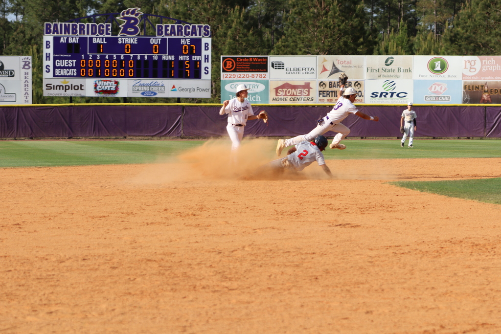 High school baseball game featuring Bainbridge Bearcats players in white uniforms fielding, pitching, catching, and running bases on a dirt and grass field, with a scoreboard, outfield fence signage, and spectators in the background.