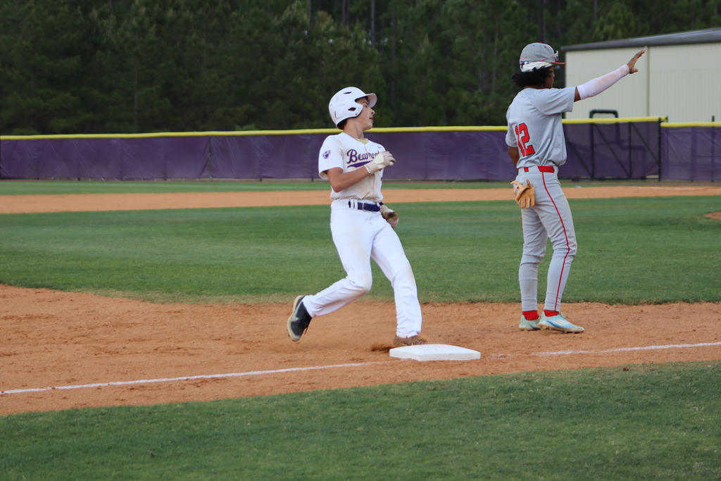 High school baseball game featuring Bainbridge Bearcats players in white uniforms fielding, pitching, catching, and running bases on a dirt and grass field, with a scoreboard, outfield fence signage, and spectators in the background.