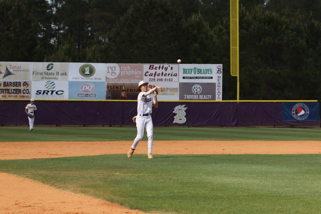 High school baseball game featuring Bainbridge Bearcats players in white uniforms fielding, pitching, catching, and running bases on a dirt and grass field, with a scoreboard, outfield fence signage, and spectators in the background.