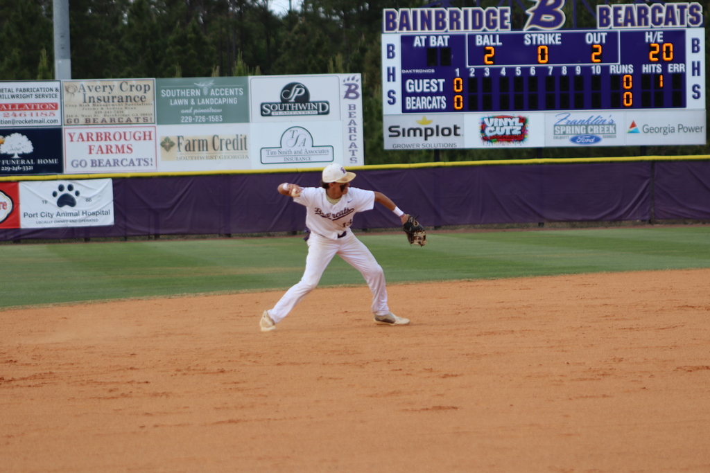 High school baseball game featuring Bainbridge Bearcats players in white uniforms fielding, pitching, catching, and running bases on a dirt and grass field, with a scoreboard, outfield fence signage, and spectators in the background.