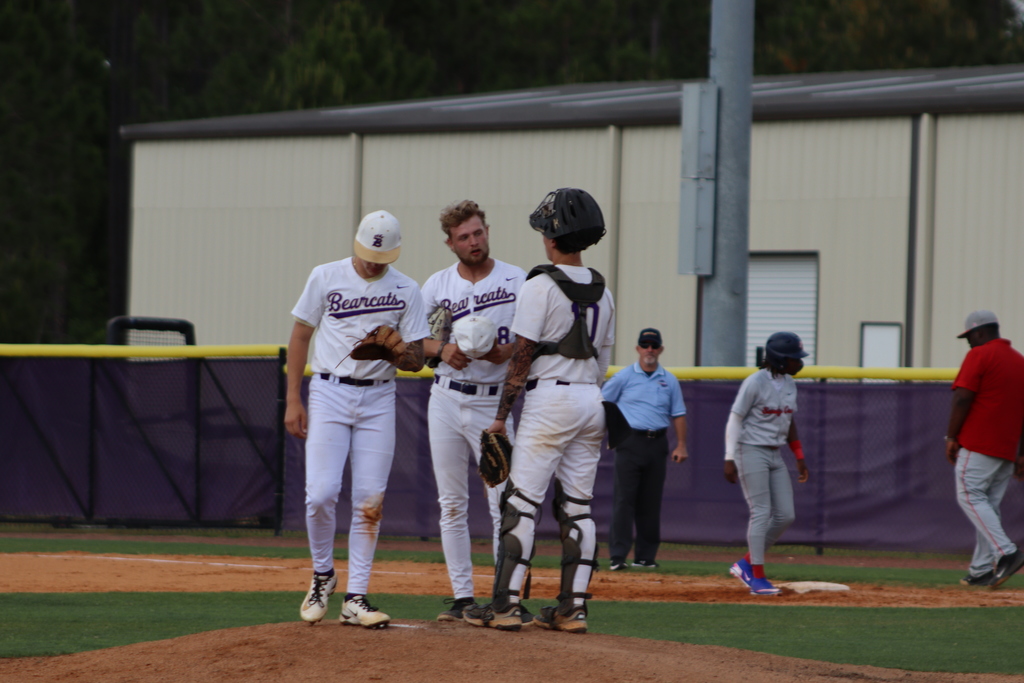 High school baseball game featuring Bainbridge Bearcats players in white uniforms fielding, pitching, catching, and running bases on a dirt and grass field, with a scoreboard, outfield fence signage, and spectators in the background.