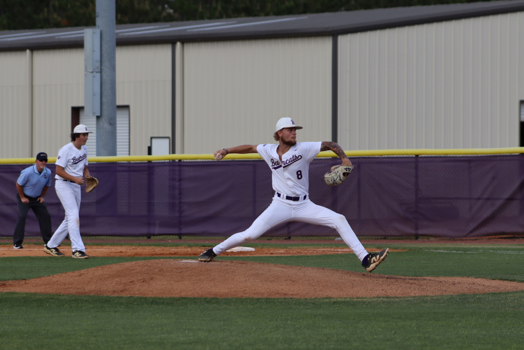 High school baseball game featuring Bainbridge Bearcats players in white uniforms fielding, pitching, catching, and running bases on a dirt and grass field, with a scoreboard, outfield fence signage, and spectators in the background.