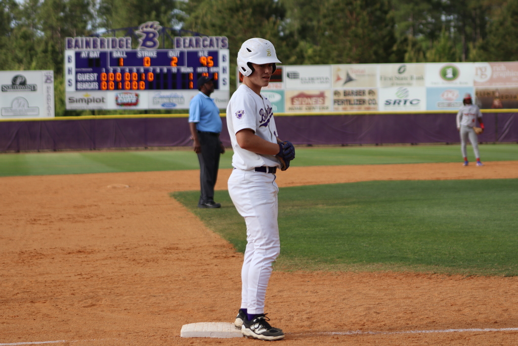 High school baseball game featuring Bainbridge Bearcats players in white uniforms fielding, pitching, catching, and running bases on a dirt and grass field, with a scoreboard, outfield fence signage, and spectators in the background.