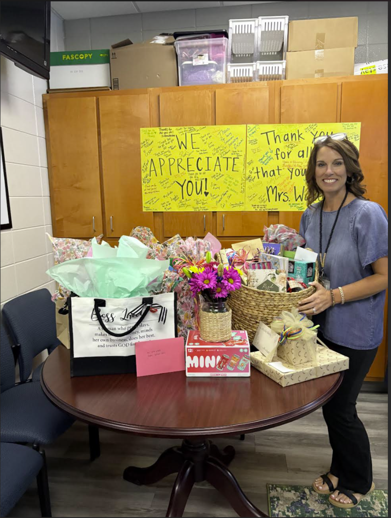 Woman smiling beside a table full of gifts and flowers with “We Appreciate You” signs behind her.