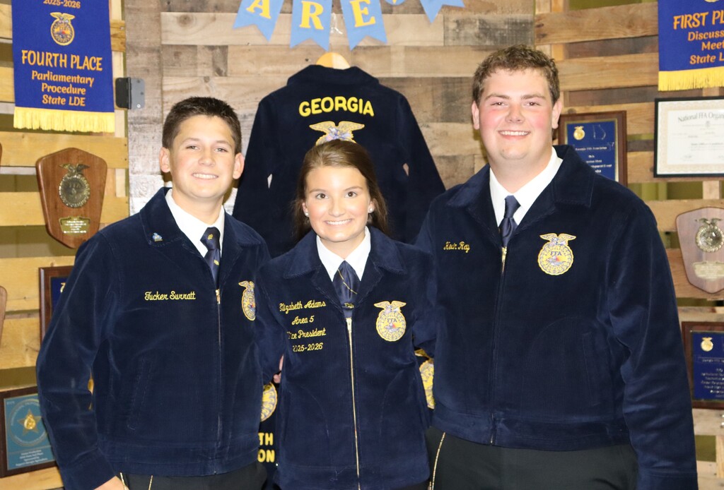 Three Bainbridge FFA members pose in official jackets in front of an awards display at the 2025–2026 FFA Banquet.