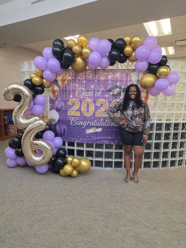 Aesha Worthy stands smiling beside a large purple, gold, and black balloon arch and graduation backdrop that reads “Class of 2025 Congratulations” during the Bearcat Stroll celebration at Jones Wheat Primary.