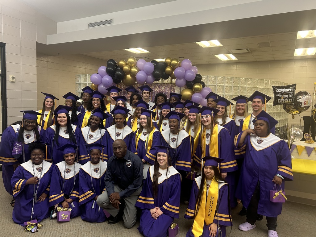A group of graduating students in purple caps and gowns pose together in a classroom setting.