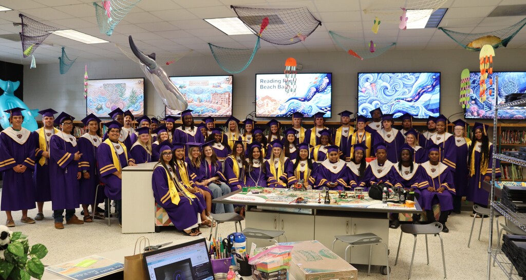 A group of graduating students in purple caps and gowns pose together in a classroom setting.